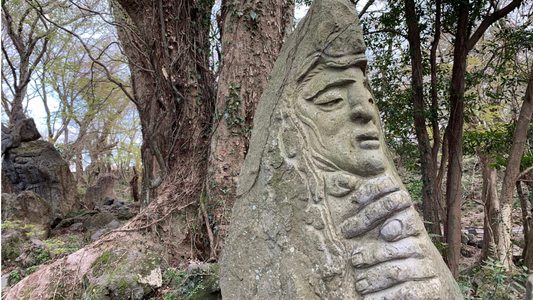 A carved stone statue with a serene face and intricate hand details, set against a backdrop of trees and nature