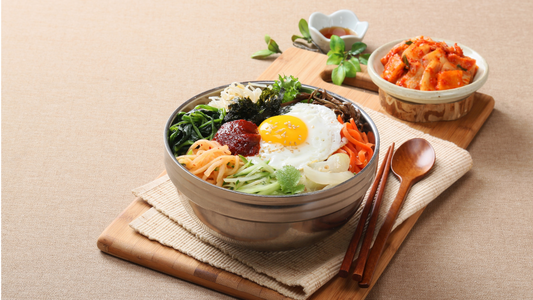 A beautifully presented bibimbap in a stone bowl, featuring a fried egg, various vegetables, and gochujang sauce, served with chopsticks, a spoon, and side dishes including kimchi on a wooden board.